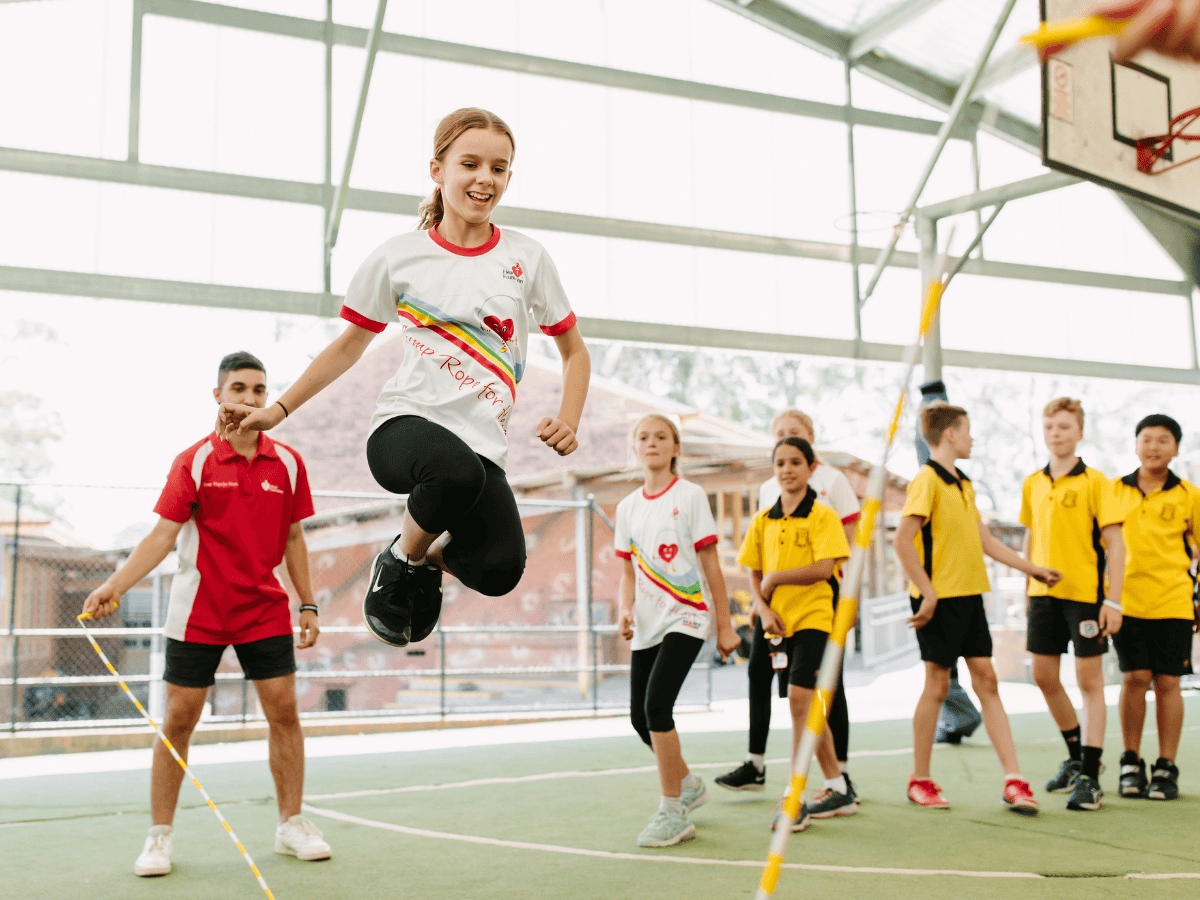 A young girl jumping over a long skipping rope surrounded by her primary school peers