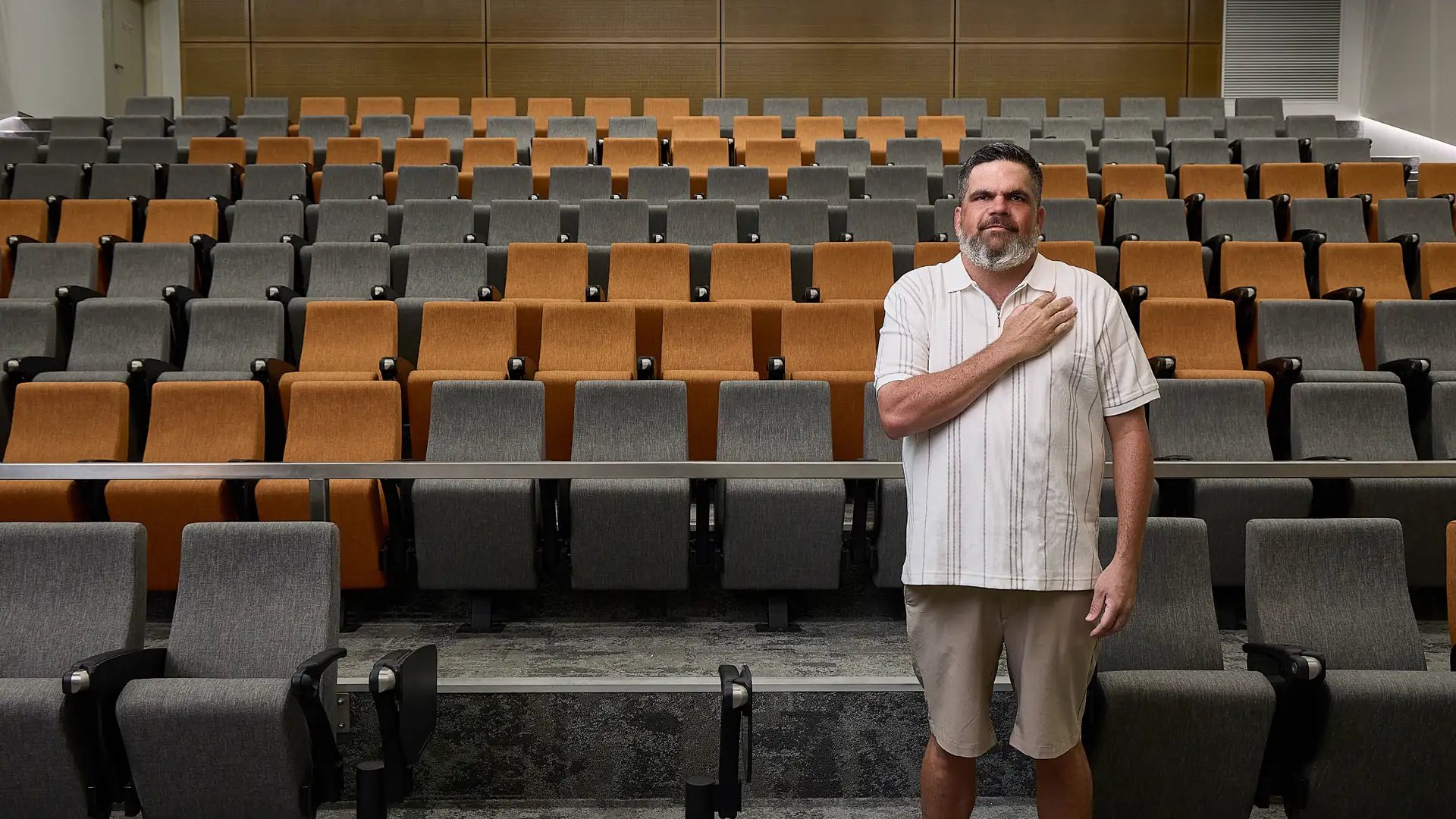 First Nations man Kieran Bush standing in an empty lecture theater with his hand placed over his heart
