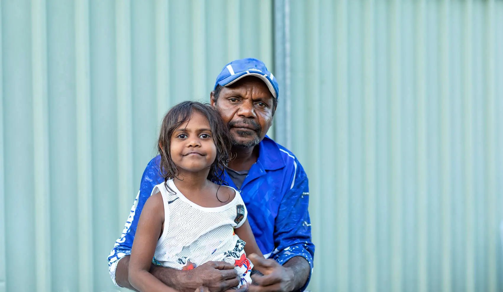 Man with arms around little kid sitting on his knee in front of tin fence