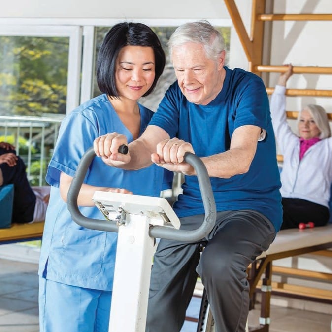 Older adult using an exercise bike with support from a healthcare professional during a rehabilitation session