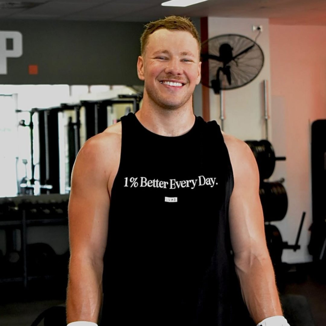 Josh, who lives with hypertrophic cardiomyopathy, is smiling at the gym, with a tank top with slogan '1% Better Every Day'