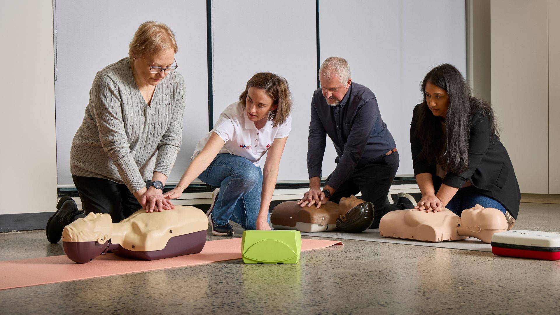 Four people kneeling and performing CPR on training mannequins during a Heart of the Nation session, with a defibrillator visible in the foreground.