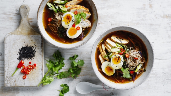 Two bowls of ramen on a table with some coriander on the side