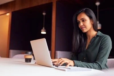 Woman sitting at table with laptop typing and coffee nearby