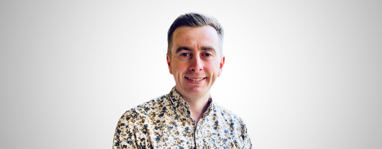 A man wearing a floral shirt smiles warmly at the camera, radiating a sense of joy and positivity.
