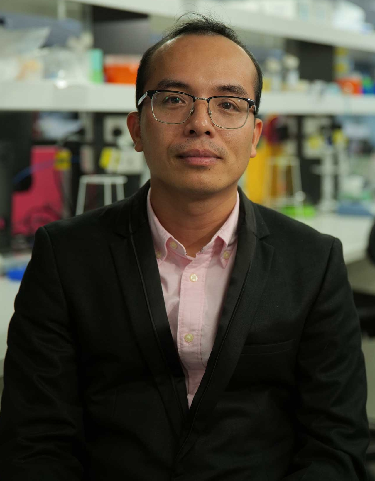 Dr Thanh Nho Do, wearing glasses and a pink shirt, black blazer, lab environment in the background