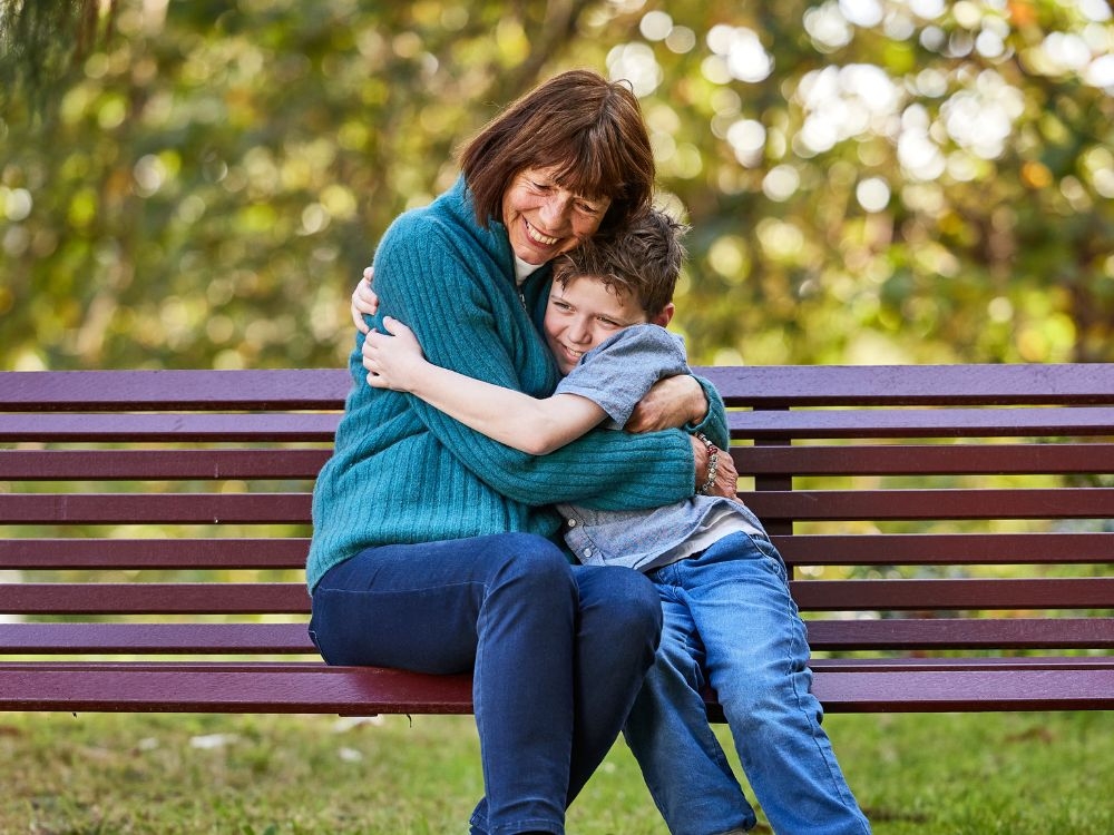 An older woman sitting on a park bench hugging her young grandson