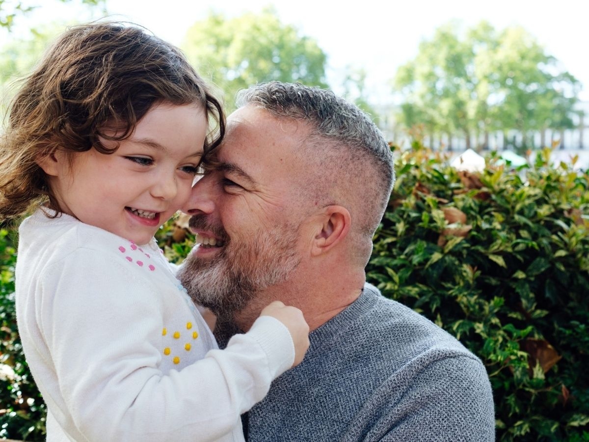 A middle-aged man holding his young daughter standing in front of a leafy hedge