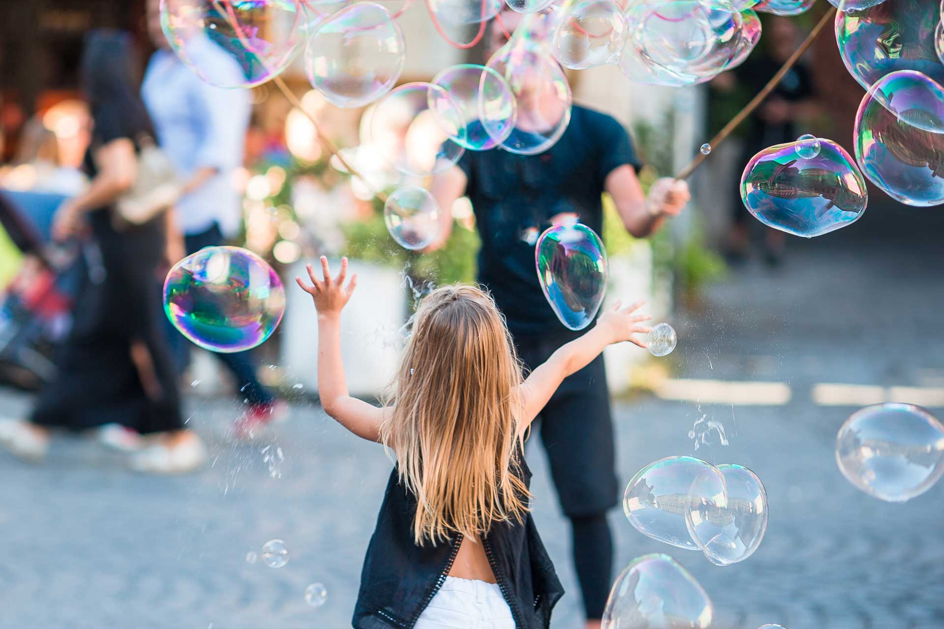 Blond haired child reaching up joyfully toward floating soap bubbles in a lively outdoor setting