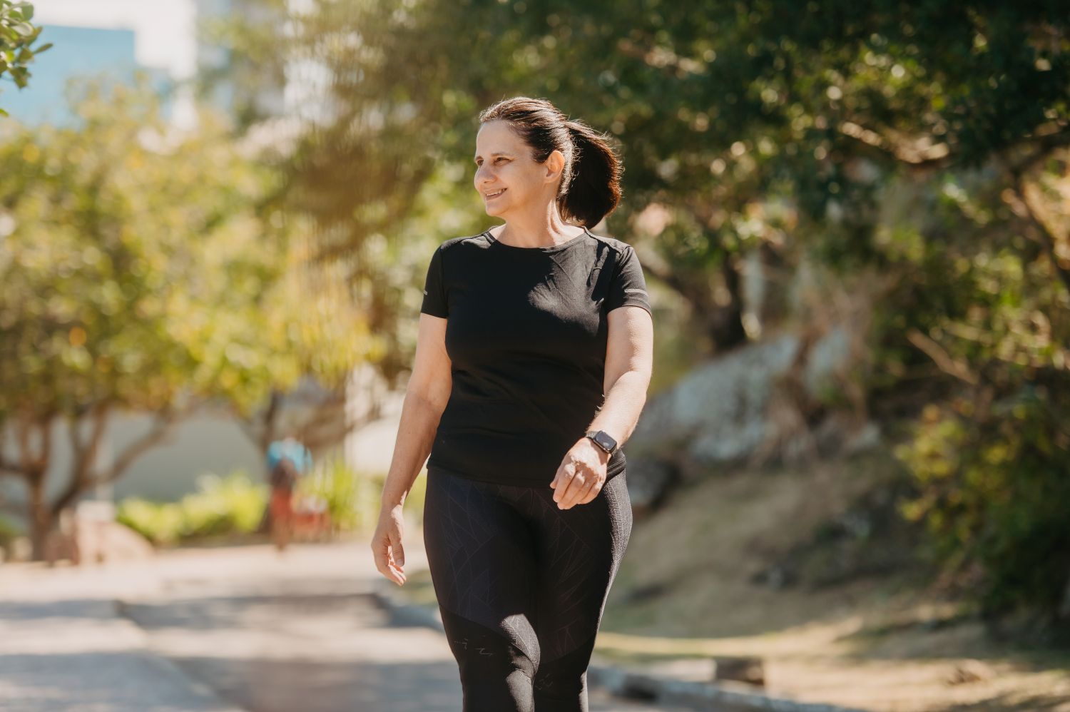 Person in black activewear walking along a sunlit, tree-lined path outdoors.