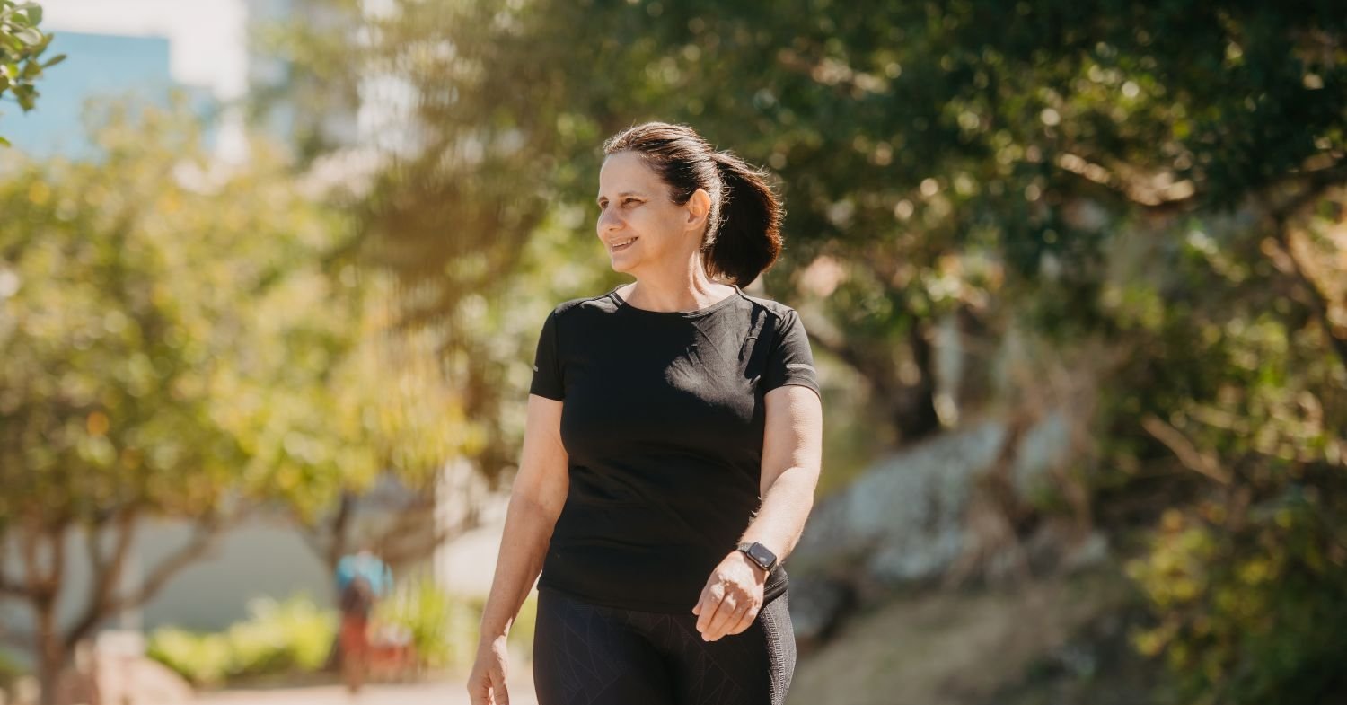Person in black activewear walking along a sunlit, tree-lined path outdoors.