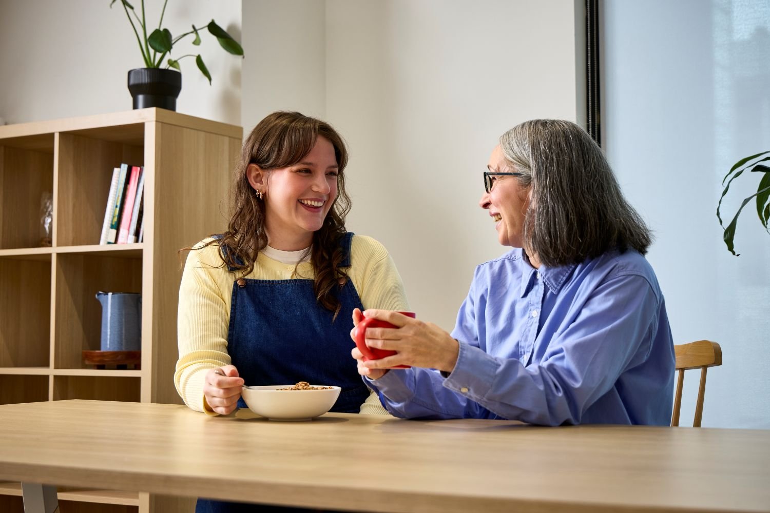 Two people sitting at a table indoors, smiling and talking while sharing a cup of tea and a bowl of cereal, with a plant and bookshelf in the background.