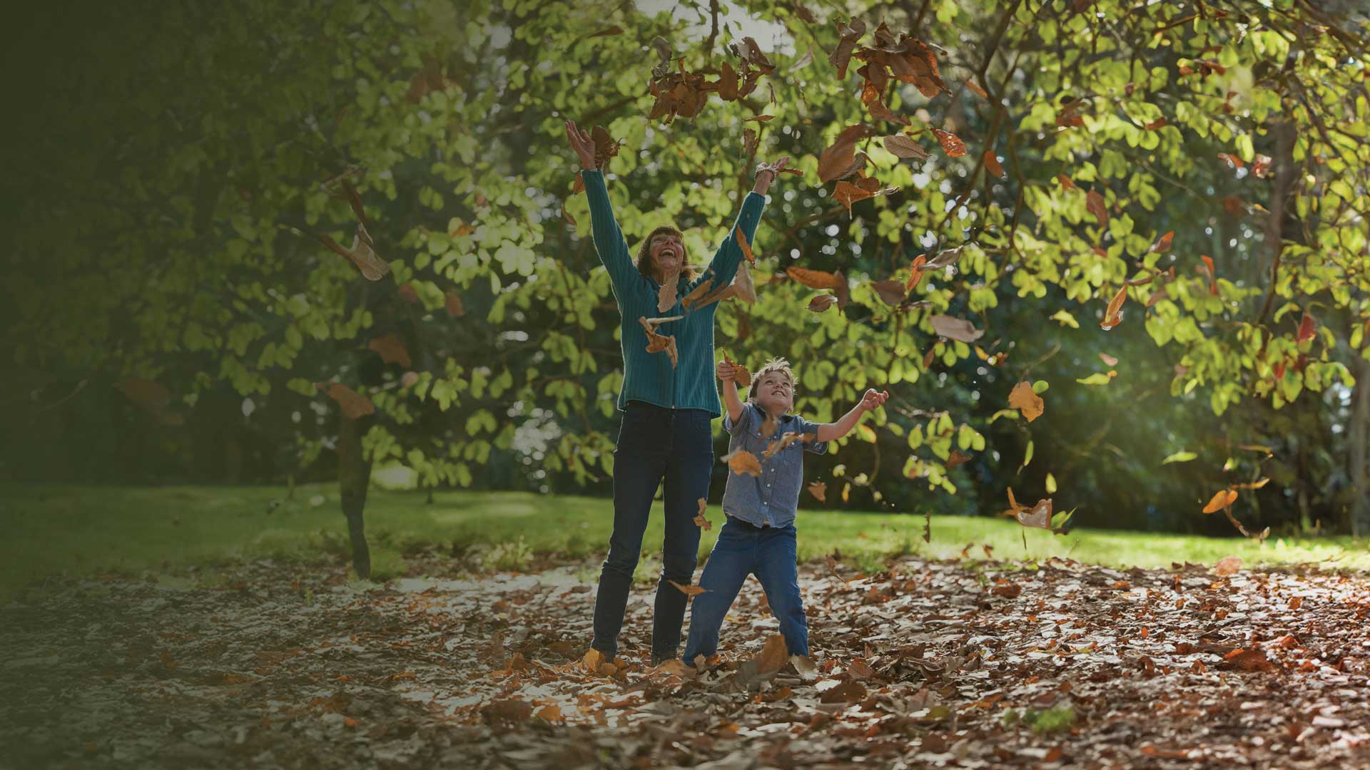 A woman and a young boy playing, throwing leaves under trees in a park