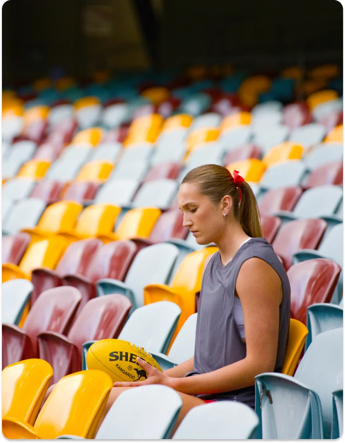 AFLW player Tay Smith sitting in the empty stands of the Gabba looking down at a football