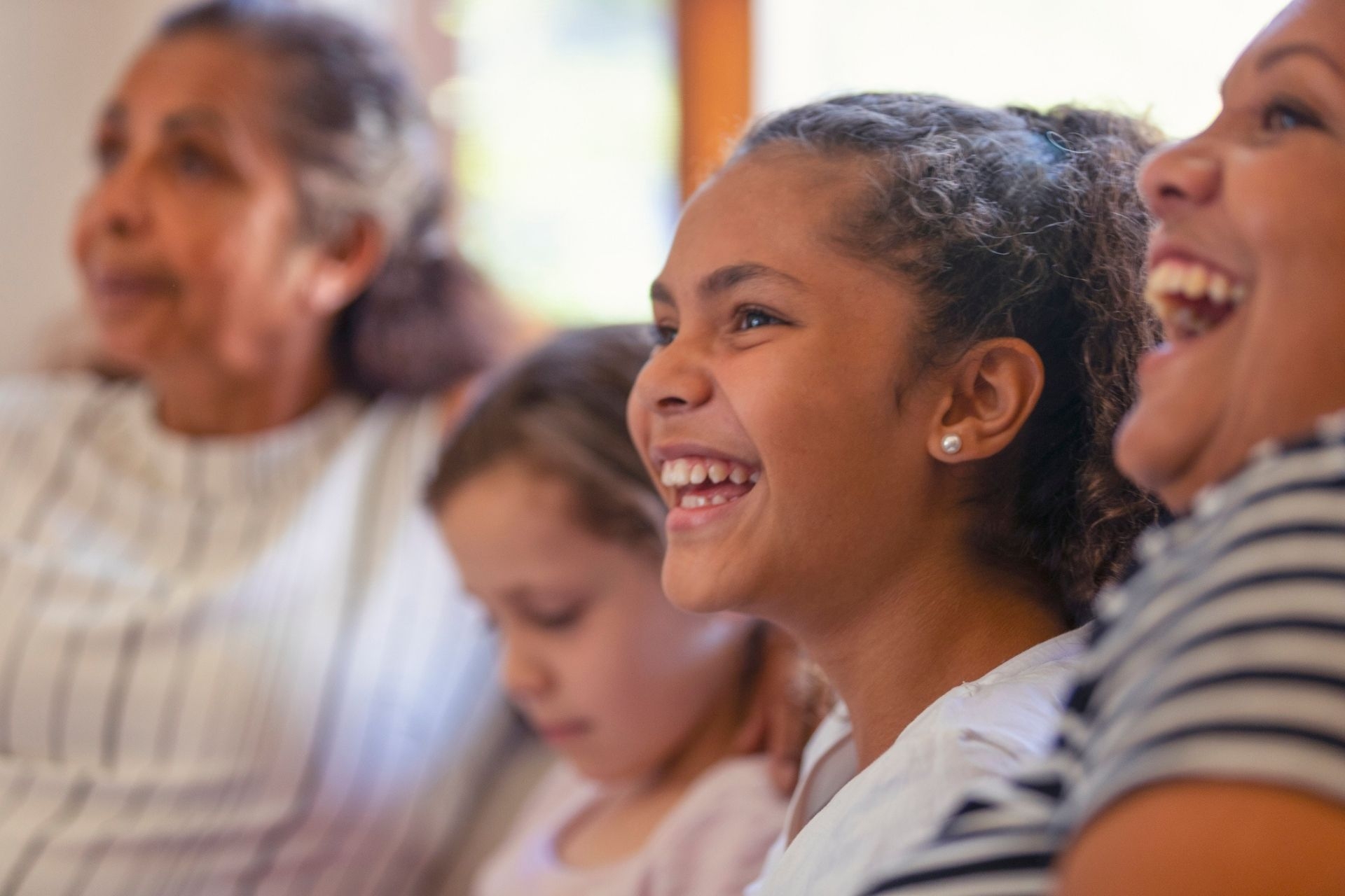 Multi generational women on the sofa. The young girls are sisters. One is a teenager on is younger. There is also a mother and grandmother. They are all happy and smiling. They are all Australian Aboriginals.