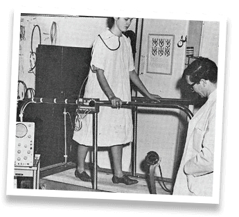 Black & white photo of a woman walking on a treadmill being observed by doctor