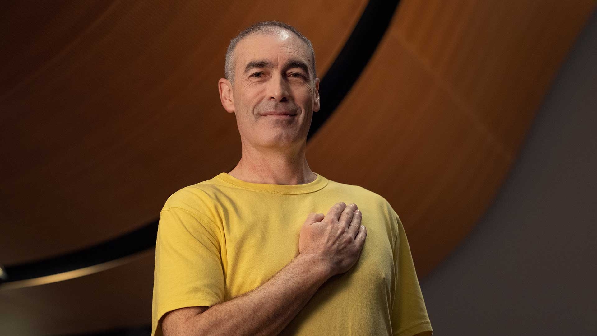 Greg Page in a yellow t-shirt with his hand on heart