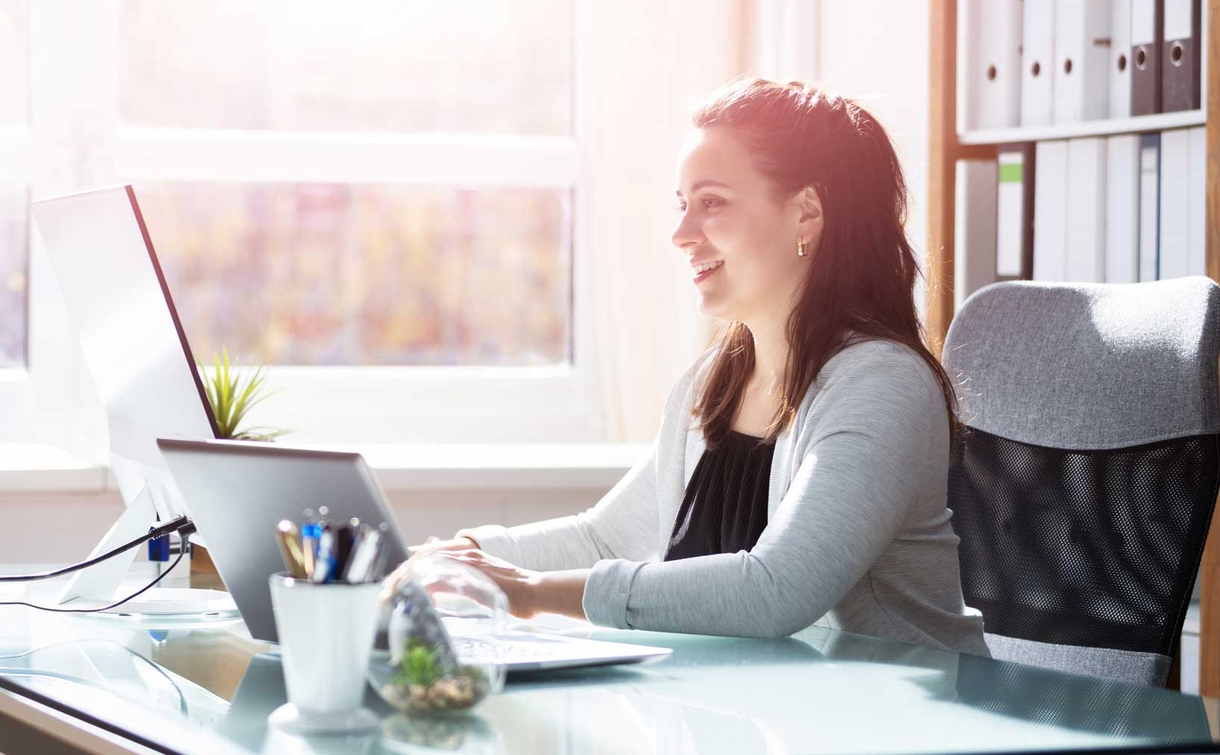 a smiling woman sitting at desk typing on laptop by a window with light streaming in