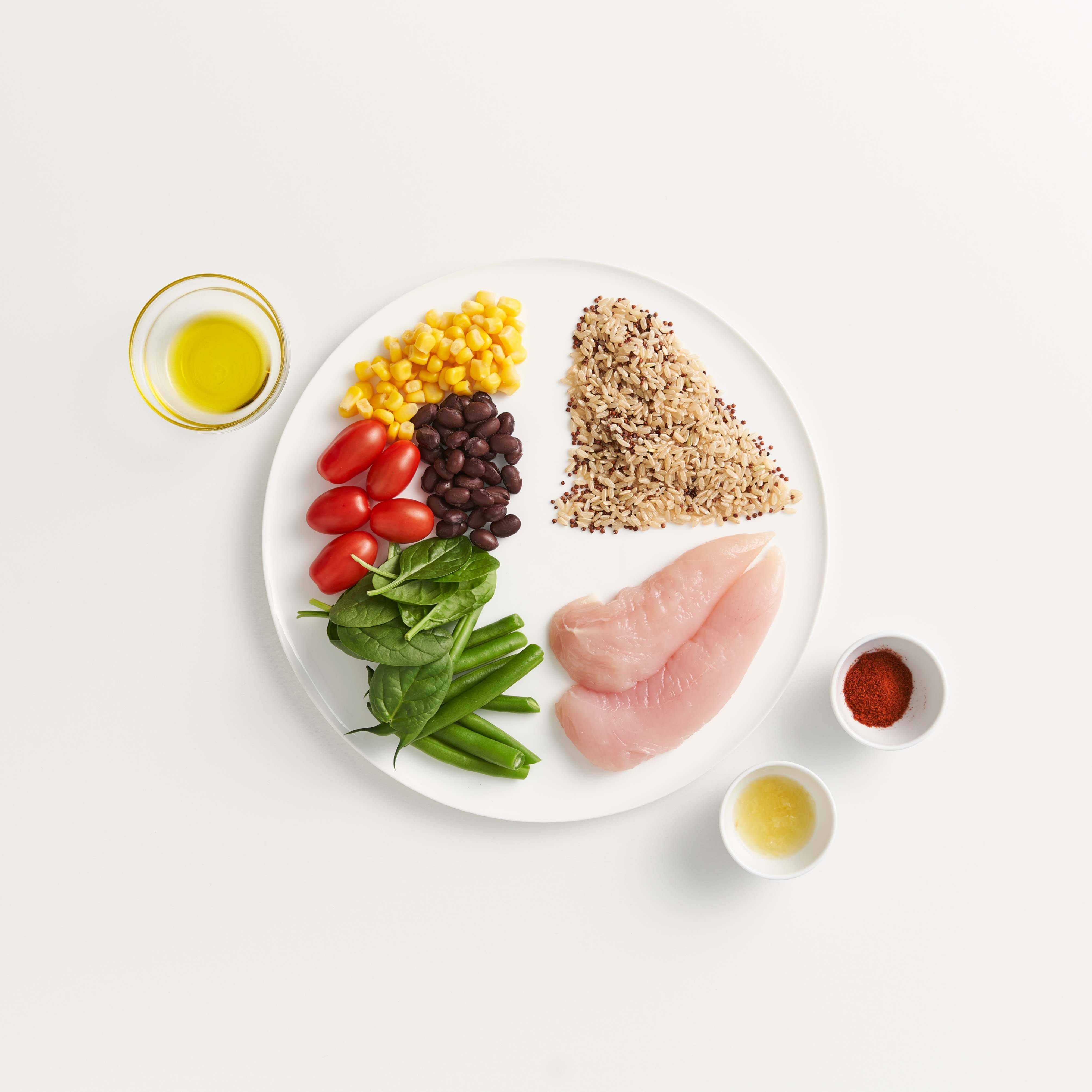 A heart-healthy eating plate with grilled chicken, spinach, cherry tomatoes, corn, and rice, surrounded by small bowls of oil, herbs, and nuts. 