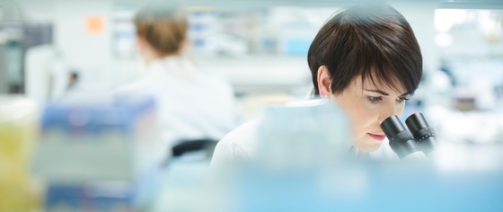 A female scientist in a lab coat examining a specimen under a microscope.