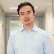Associate Professor Quan Huynh, in a light blue shirt, stands in the middle of a hallway lined with office doors and white walls.