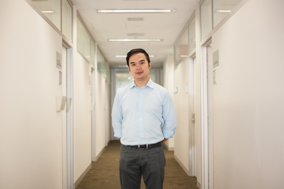 Associate Professor Quan Huynh, in a light blue shirt, stands in the middle of a hallway lined with office doors and white walls.