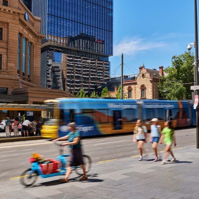 People walking and cycling along a city street with a tram travelling through the background.