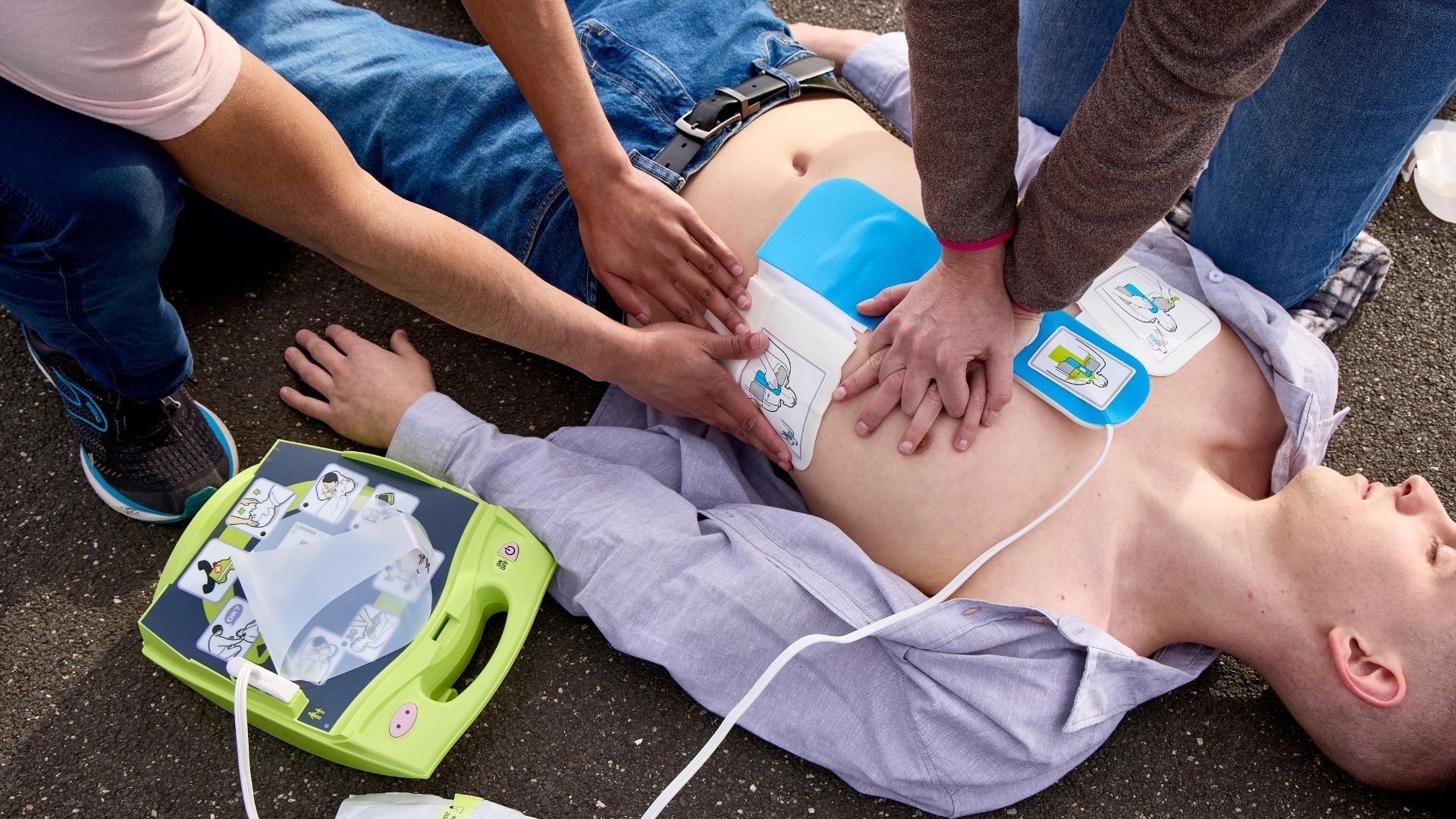Two individuals performing CPR on a person lying on the ground. One is giving chest compressions while the other applies AED pads to the person's chest. The AED device is visible beside them.