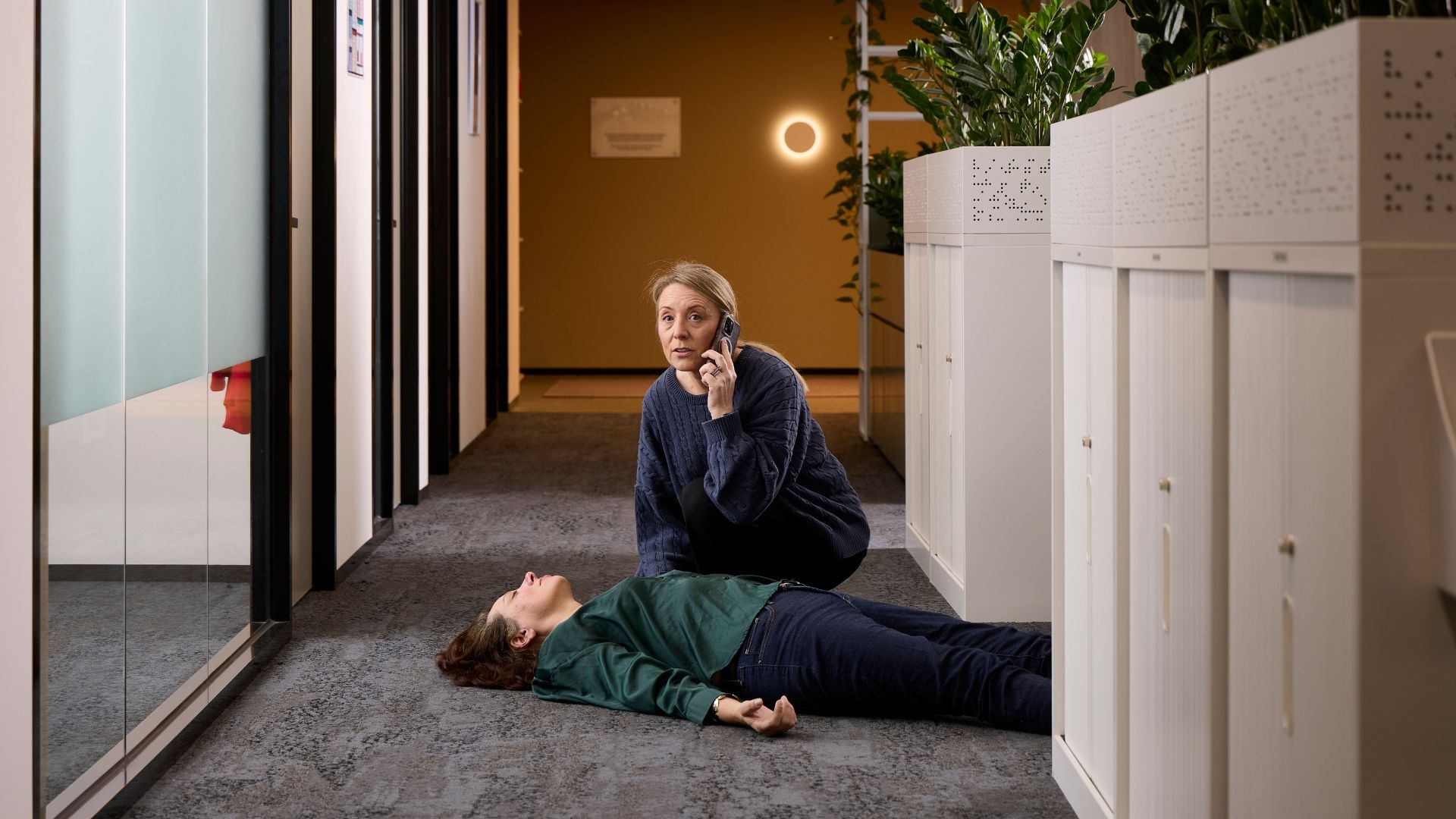 A woman kneeling on the phone to Triple Zero, next to a woman who is cardiac arrest