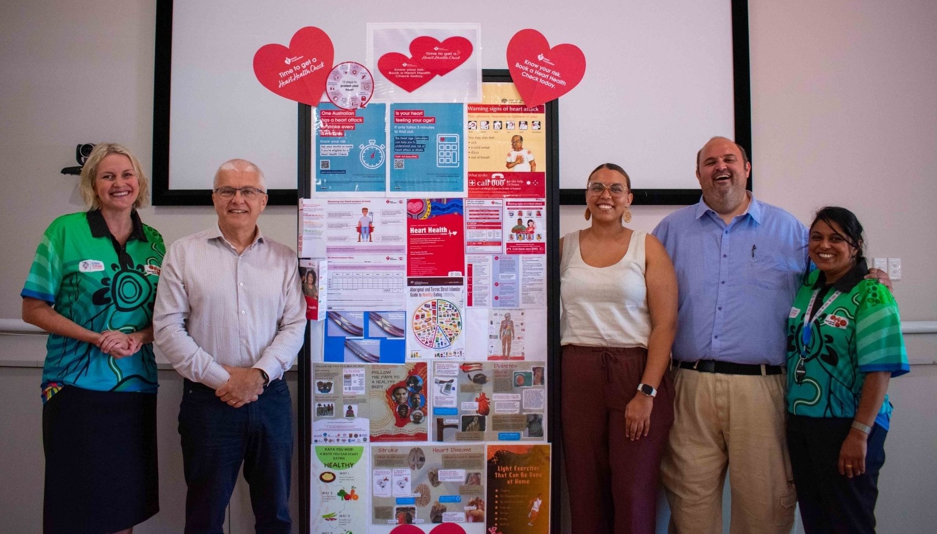 A group of Heart Foundation and Derbarl Yerrigan Health Service standing next to a pinboard of community health messages. 