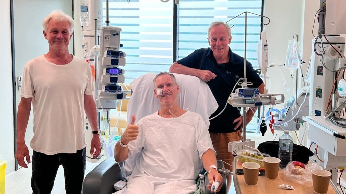 A patient recovering in hospital gives a thumbs up while seated in a chair, alongside two visitors in a hospital room.