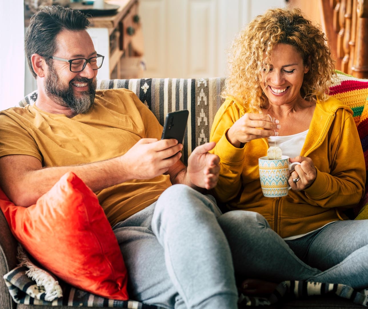 A woman drinking a cup of tea whilst a man is on his phone, they are both laughing and enjoying each other company.