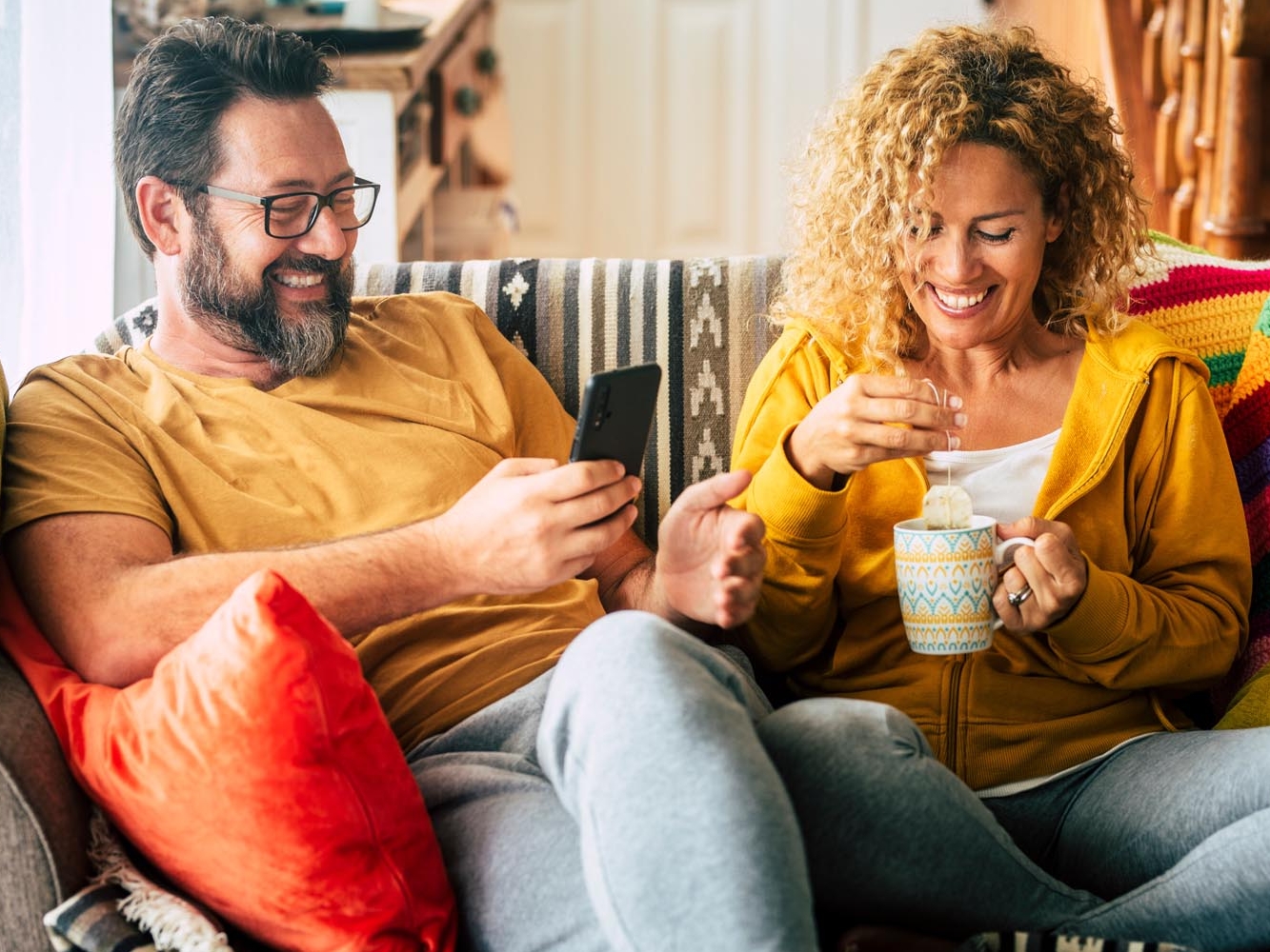 Man and woman seated on a couch together laughing, the man is also interacting on his phone