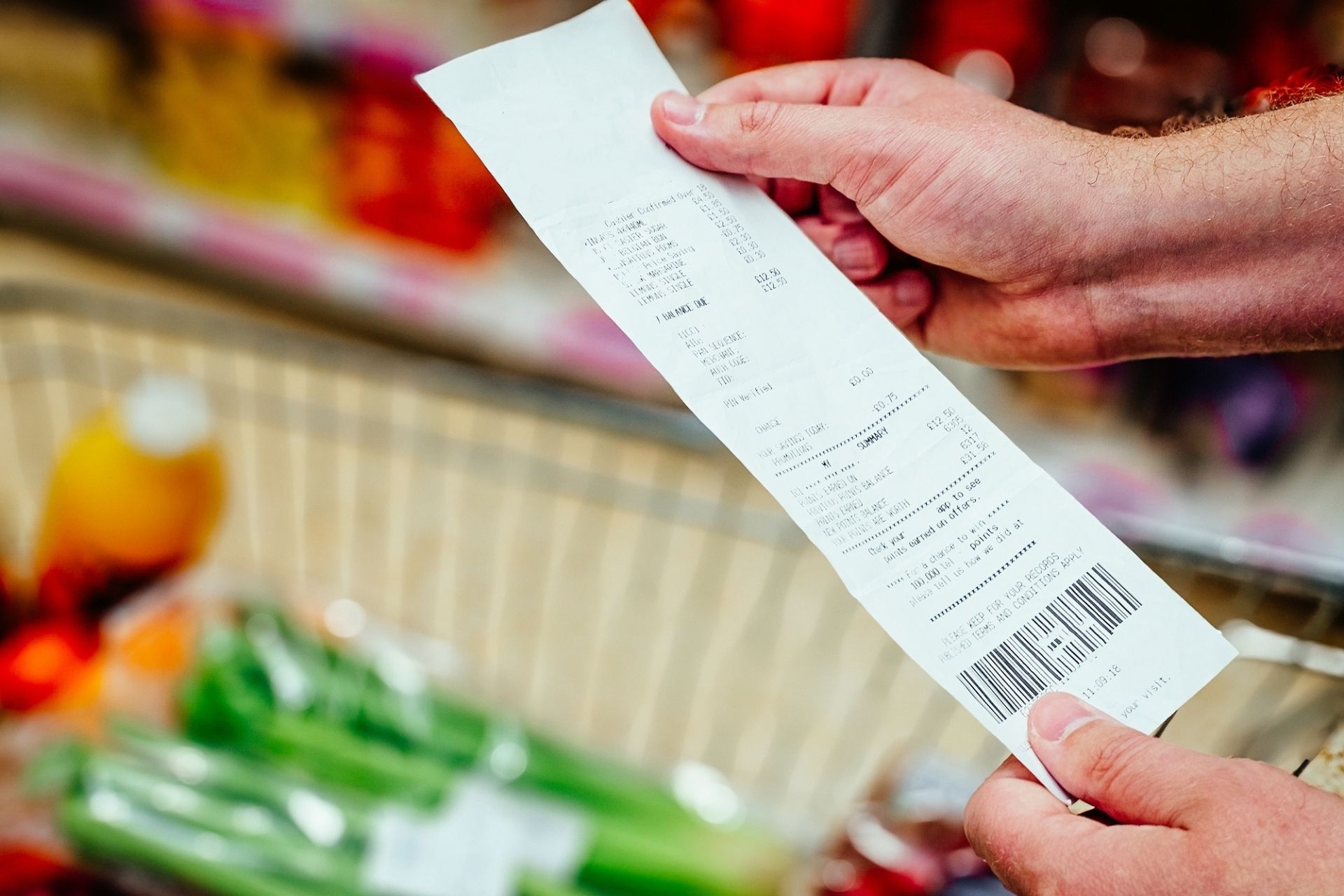 Close up color image depicting a man's hands holding and checking his groceries bill in the supermarket after making his purchases at the checkout. Focus on the receipt with the contents of the shopping trolley defocused in the background.