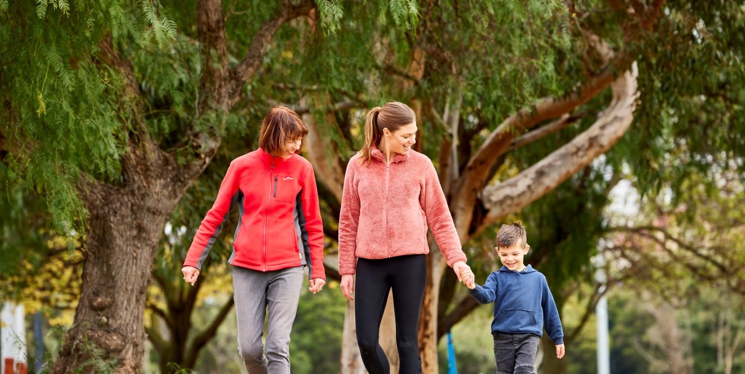 Three people walking together along a gravel path in a park, holding hands, with large trees and grass on either side.