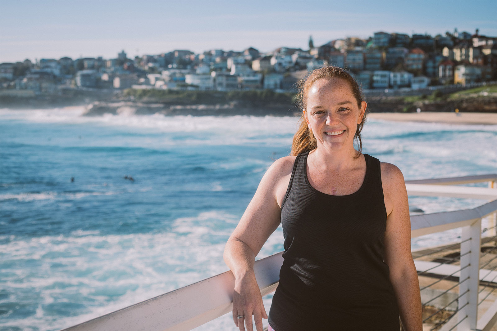 A woman in a black tank top stands on a boardwalk, gazing at the vast ocean ahead.