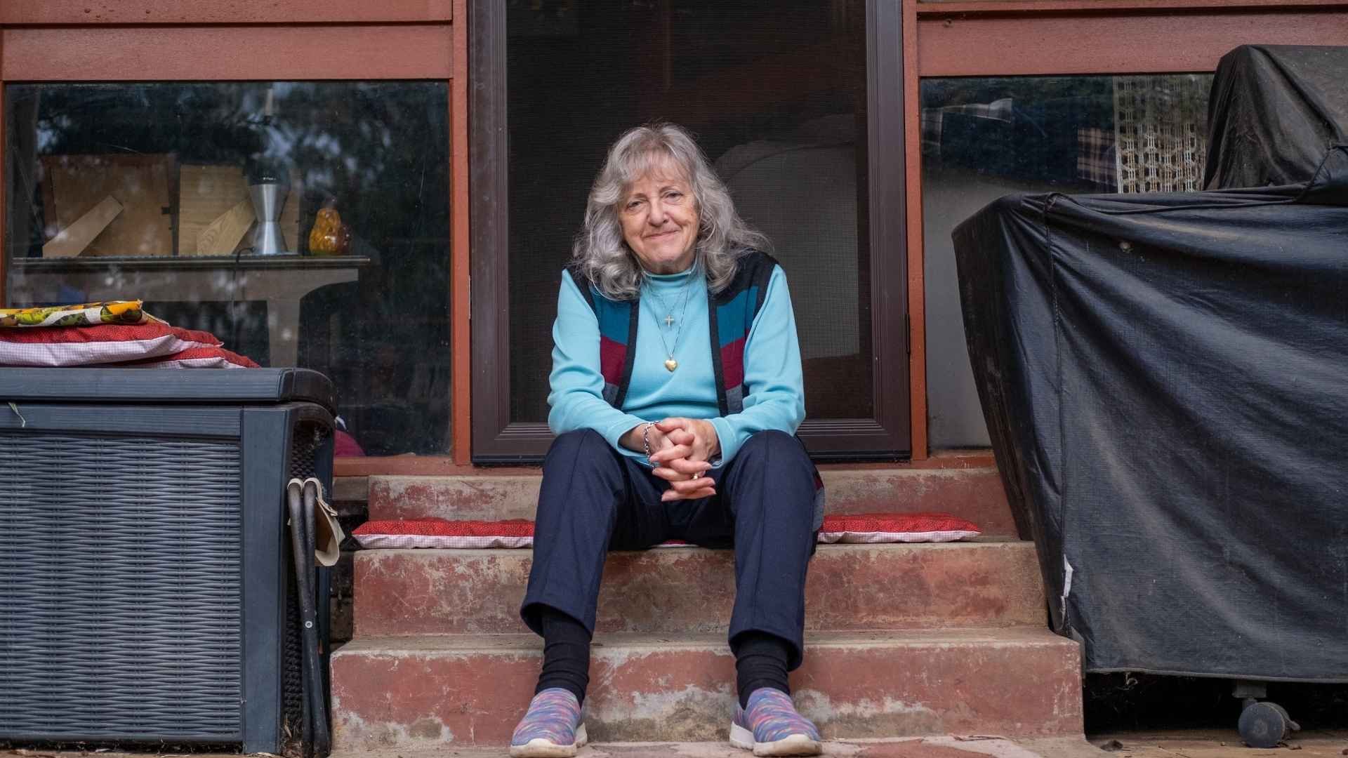 Woman with grey hair in blue top smiling, sitting on stairs at the back door of her house