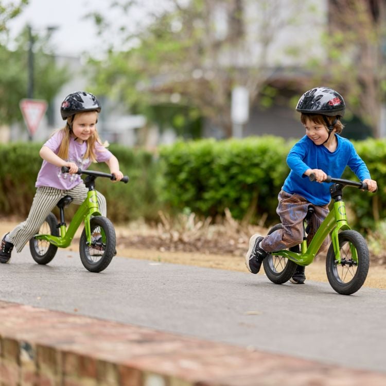 Two children riding balance bikes on a paved path in a park