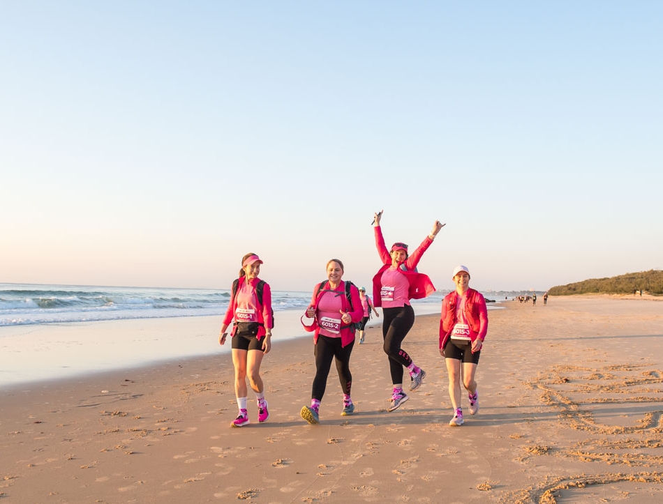 Four Coastrek participants walking on a beautiful beach, one is jumping in the air