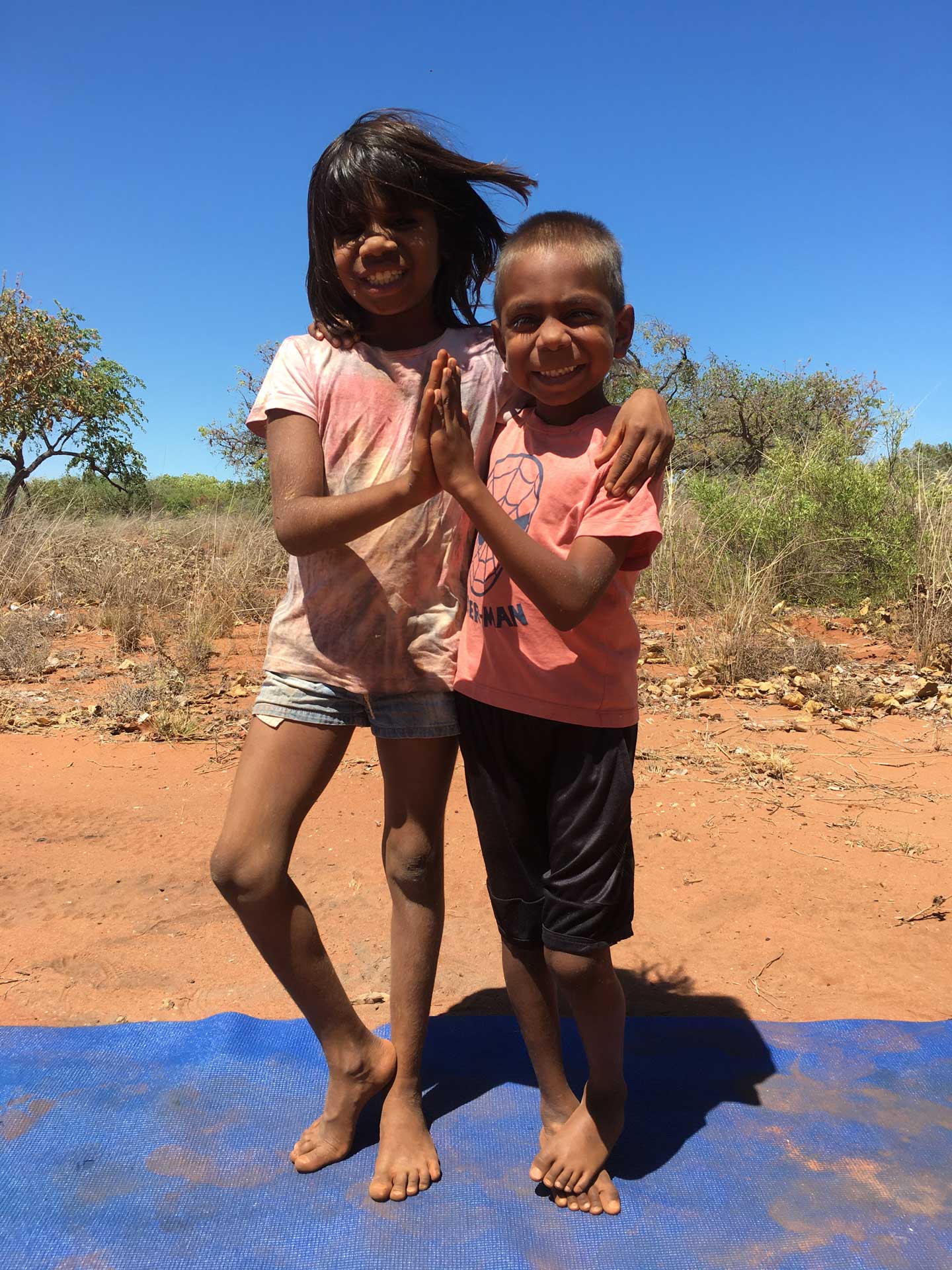 Two First Nations kids holding their hands together set against a red desert / rural backdrop 