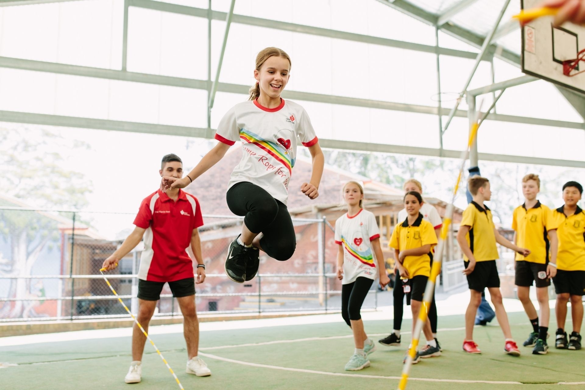 A young girl jumping over a long skipping rope surrounded by her classmates