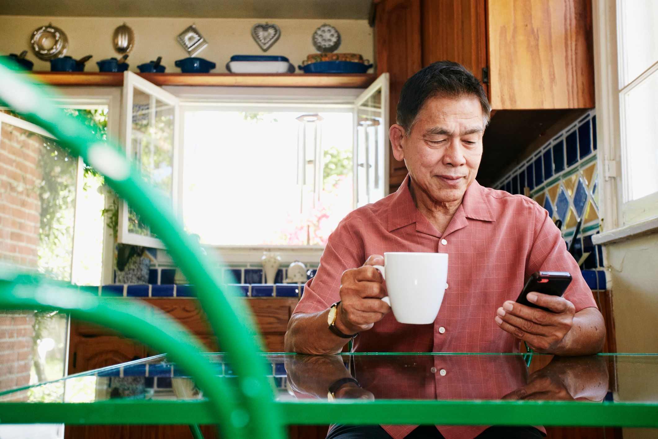 An older man sitting at his kitchen table, looking down at his smartphone in one hand and holding a mug in the other. 