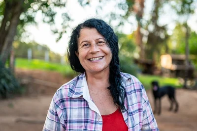 Woman smiling with dark wavy hair, check plaid shirt, standing in outside near tree and grass