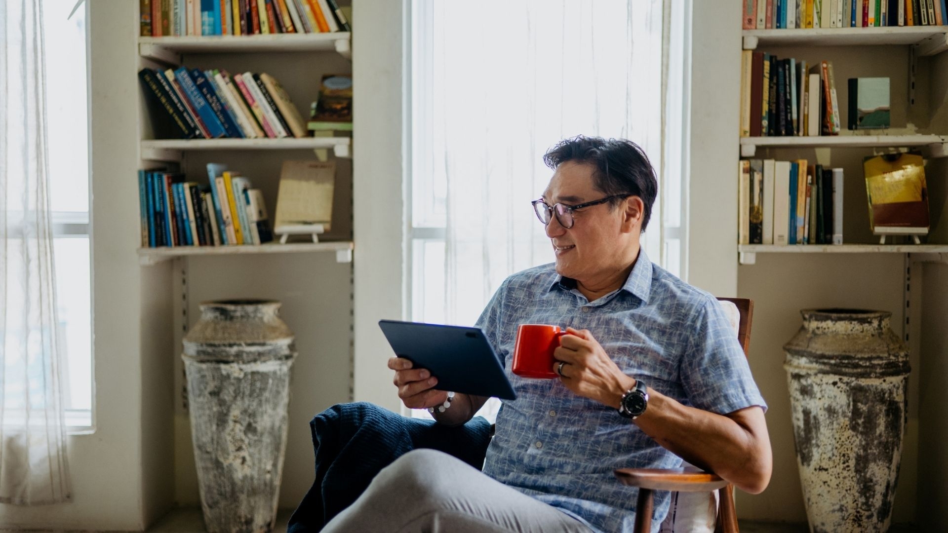 A person sits in a chair holding a red mug and a tablet, wearing glasses and a short-sleeved shirt. Behind them are two large vases and bookshelves filled with books. Two windows with sheer curtains let in natural light.