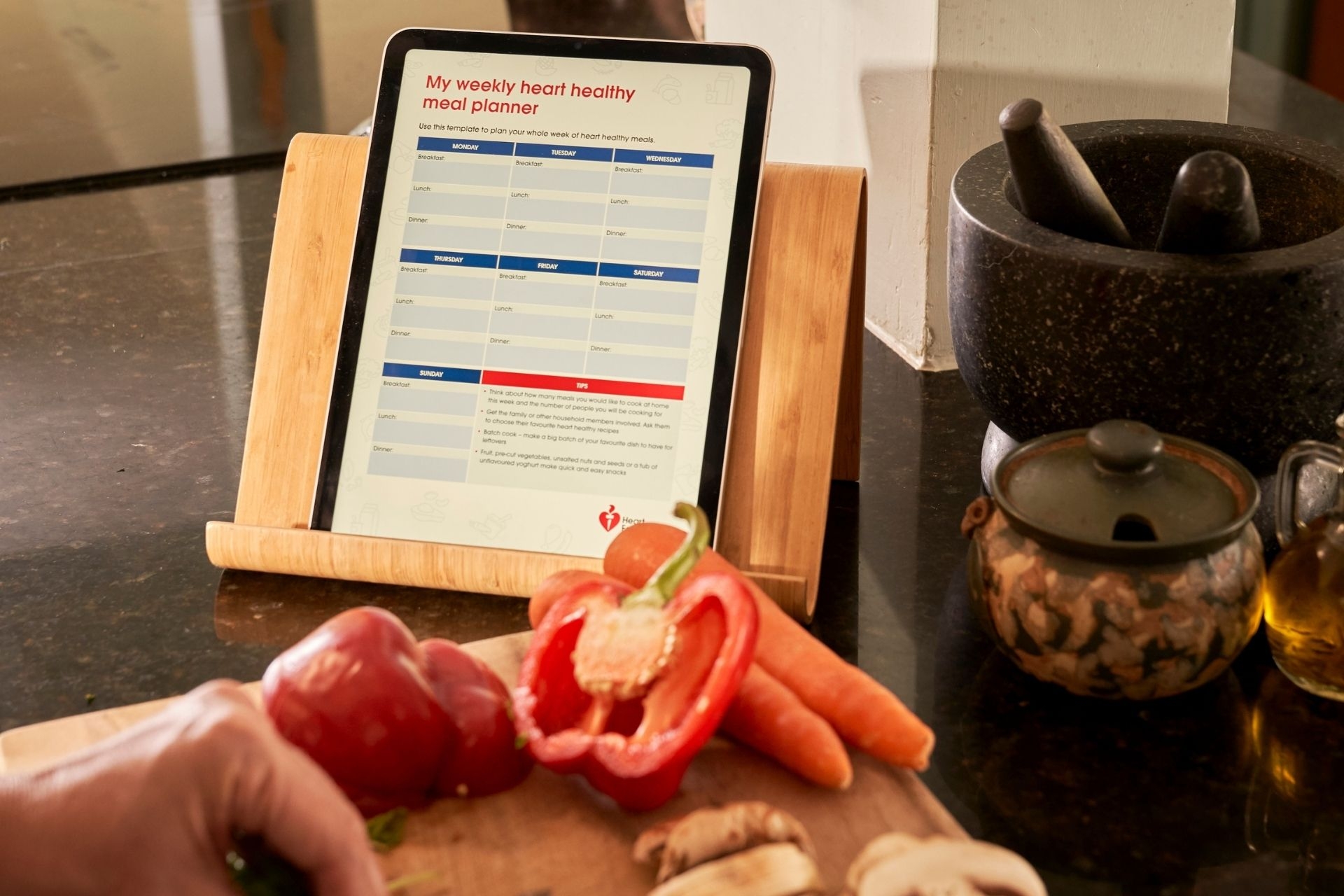 A tablet sitting on a stand on a kitchen bench, the tablet is displaying the Heart Foundation's weekly heart-healthy meal planner