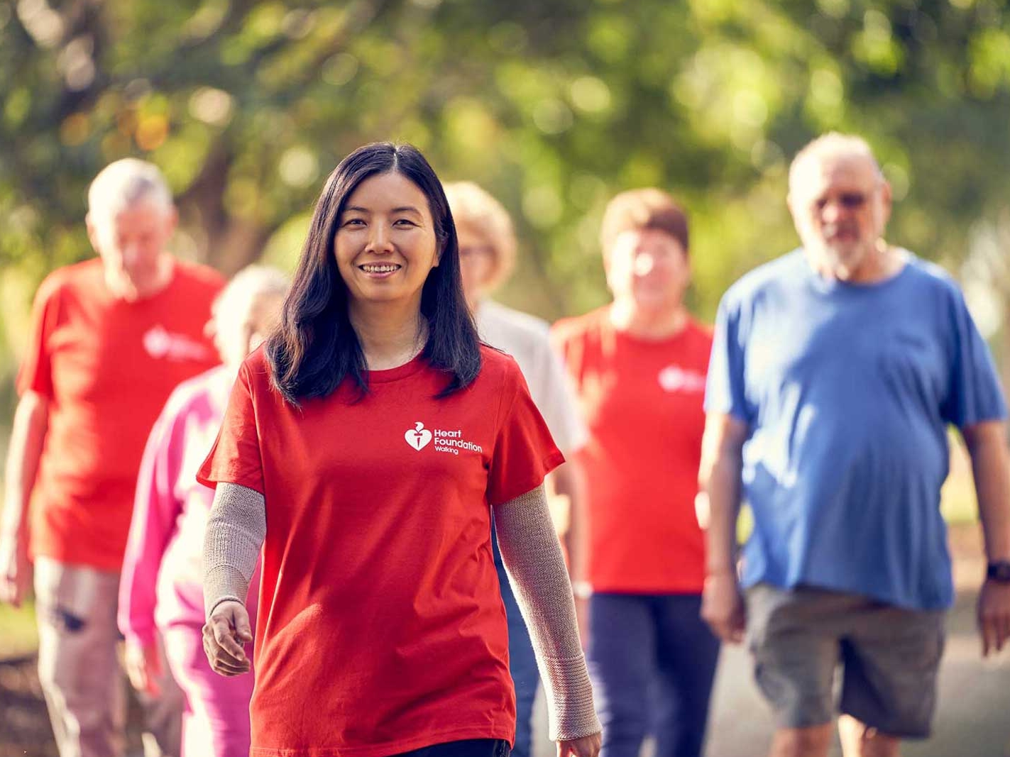 Group of people walking in a park, with a dark haired smiling young woman in red HF tshirt at the lead