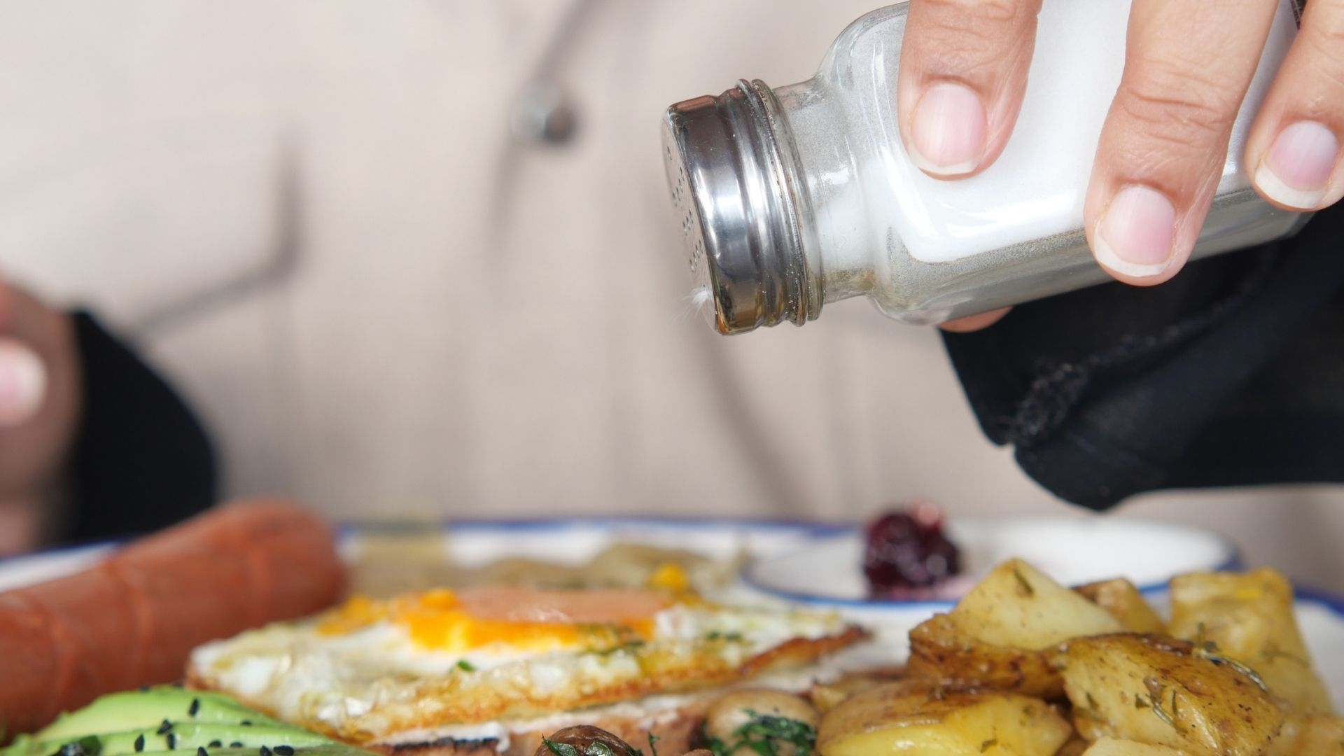 A hand adds salt to a breakfast plate filled with various foods.