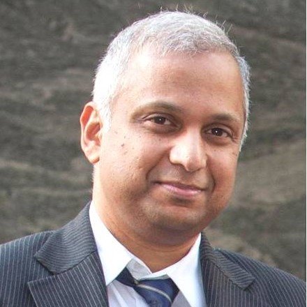 Professor Sandosh Padmanabhan in a suit and tie posing confidently in front of a plain wall.