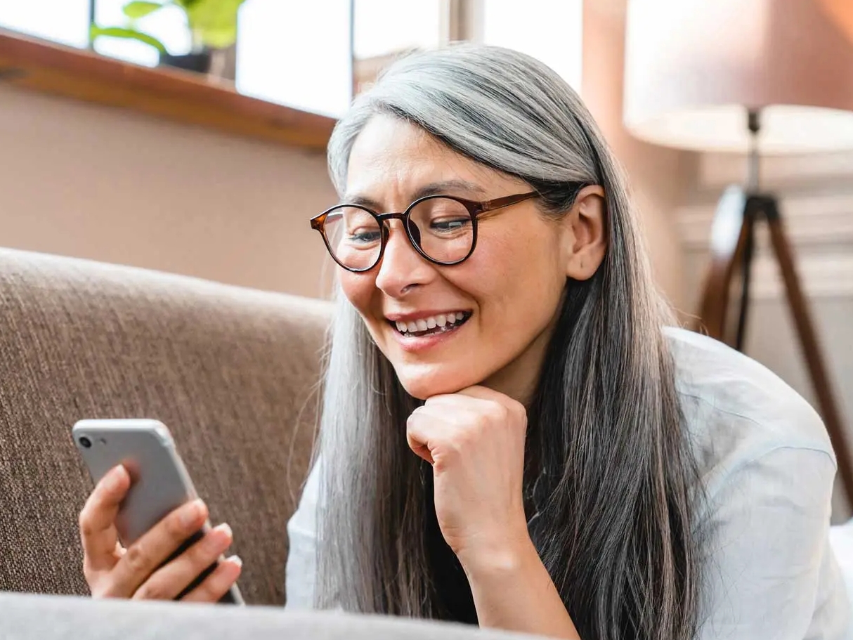 Smiling woman with glasses and long grey hair is smiling and looking at her phone
