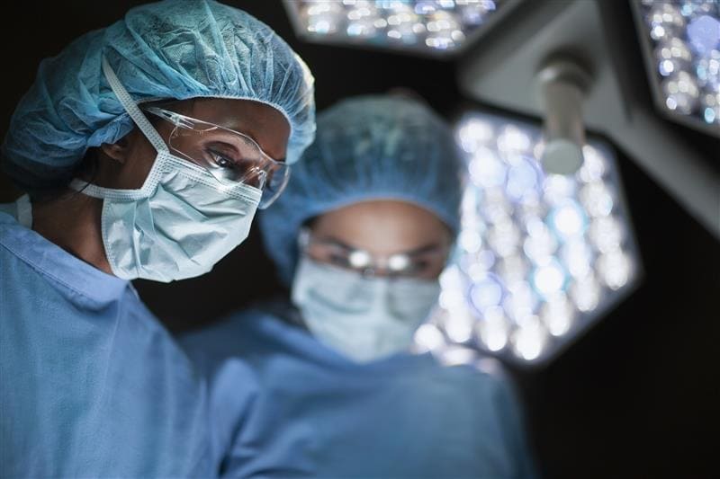 Two surgeons in blue scrubs and masks focus intently under bright surgical lights for a bypass surgery. 